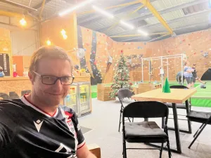 A picture of me sitting infront of a green plastic tree with shiny things on it, and a bouldering wall beyond.